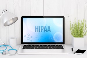 A laptop showing HIPAA word on its screen and a table lamp, vase, pen, and mobile phone are placed around it