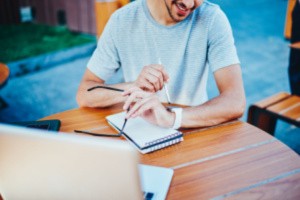 An employee checking time from his watch while working in his laptop and making notes