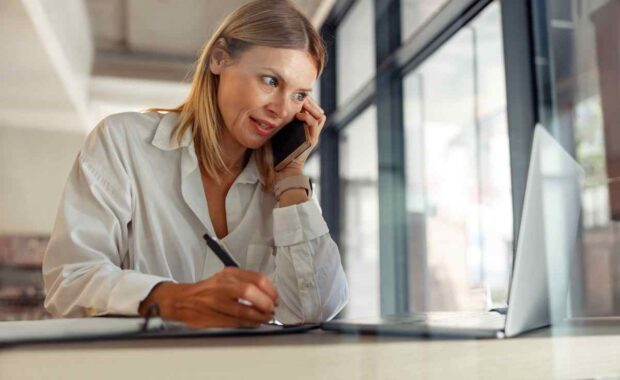 Woman entrepreneur making notes and talking by phone with client while sitting in coworking